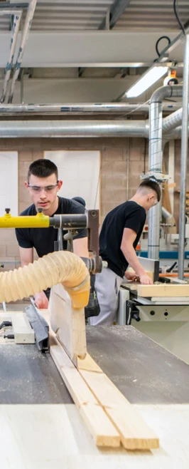 Joinery students working in a well-equipped workshop, using woodworking machinery to cut timber pieces, with other students and staff collaborating in the background. Joinery students working in a well-equipped workshop, using woodworking machinery to cut timber pieces, with other students and staff collaborating in the background.